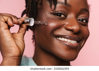 Black Shirtless Woman With Towel Smiling And Applying Face Serum Isolated Over Pink Background