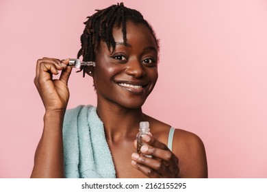 Black Shirtless Woman With Towel Smiling And Applying Face Serum Isolated Over Pink Background