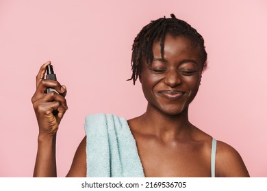 Black Shirtless Woman With Towel Smiling And Applying Face Serum Isolated Over Pink Background