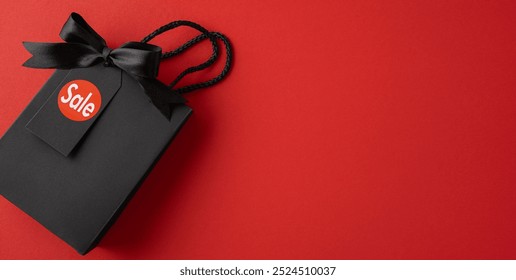 A black shopping bag with a sale tag and bow on a vibrant red background, symbolizing Black Friday shopping and present buying opportunities - Powered by Shutterstock