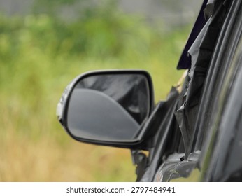 A black side view mirror attached to a damaged car door with torn window tint captured in a close up shot