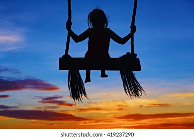 Black Silhouette Of Baby Girl Flying High With Fun On Rope Swing On Blue Orange Sunset Sky Background. 