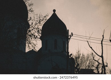  A black silhouette of a church steeple with a crow perched on top. The church is surrounded by trees with bare branches, and the sky is dark in the evening - Powered by Shutterstock