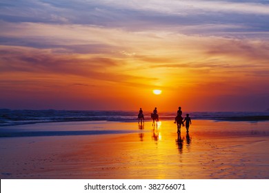 Black silhouette of happy family having horse riding adventure on sand sea beach on background of sunset sky. Active parents and people outdoor activity on tropical summer vacations with children.