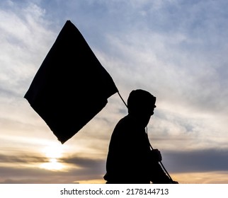black silhouette of man with flag on a sunset bright cloudy sunset evening sky, dark shadow of a rebel with black standart banner  - Shutterstock ID 2178144713