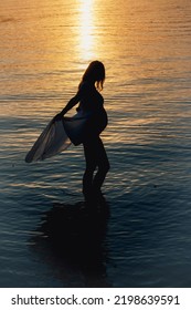 Black Silhouette Of A Pregnant Woman With A Baby Bump Or Belly Standing In The Water Of A Lake Or Sea During Romantic Sunset Or Sunrise 