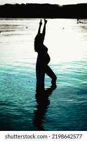 Black Silhouette Of A Pregnant Woman With A Baby Bump Or Belly Raising Her Arms In The Air To Make A Peace Or Victory Sign Like A Winner Or Champion While Standing Alone In The Water Of A Lake Or Sea 