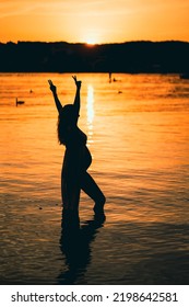 Black Silhouette Of A Pregnant Woman With A Baby Bump Or Belly Raising Her Arms In The Air To Make A Peace Or Victory Sign Like A Winner Or Champion While Standing Alone In The Water Of A Lake Or Sea 