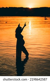 Black Silhouette Of A Pregnant Woman With A Baby Bump Or Belly Raising Her Arms In The Air To Make A Peace Or Victory Sign Like A Winner Or Champion While Standing Alone In The Water Of A Lake Or Sea 