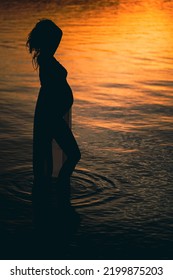 Black Silhouette Of A Sexy Beautiful Pregnant Woman With A Baby Bump Or Belly Standing In The Water Of A Lake Or Sea During Romantic Orange Sunset Or Warm Sunrise 
