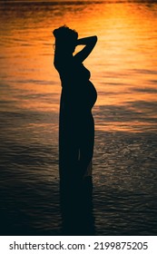 Black Silhouette Of A Sexy Beautiful Pregnant Woman With A Baby Bump Or Belly Standing In The Water Of A Lake Or Sea During Romantic Orange Sunset Or Warm Sunrise 
