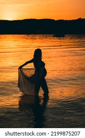 Black Silhouette Of A Sexy Beautiful Pregnant Woman With A Baby Bump Or Belly Standing In The Water Of A Lake Or Sea During Romantic Orange Sunset Or Warm Sunrise 
