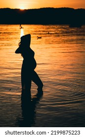 Black Silhouette Of A Sexy Beautiful Pregnant Woman With A Baby Bump Or Belly Standing In The Water Of A Lake Or Sea During Romantic Orange Sunset Or Warm Sunrise 
