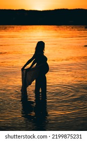 Black Silhouette Of A Sexy Beautiful Pregnant Woman With A Baby Bump Or Belly Standing In The Water Of A Lake Or Sea During Romantic Orange Sunset Or Warm Sunrise 
