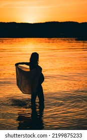 Black Silhouette Of A Sexy Beautiful Pregnant Woman With A Baby Bump Or Belly Standing In The Water Of A Lake Or Sea During Romantic Orange Sunset Or Warm Sunrise 
