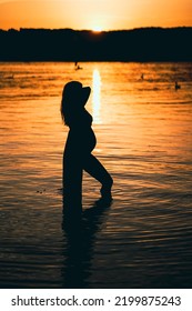 Black Silhouette Of A Sexy Beautiful Pregnant Woman With A Baby Bump Or Belly Standing In The Water Of A Lake Or Sea During Romantic Orange Sunset Or Warm Sunrise 
