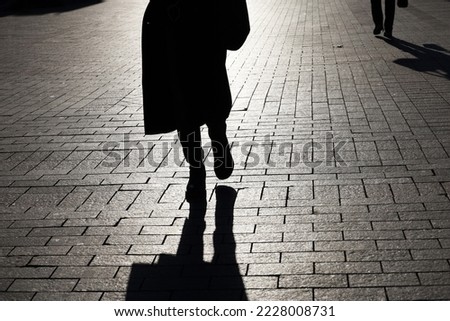Black silhouette and shadow of lonely woman walking on a street and person on background. Female legs on paved city sidewalk