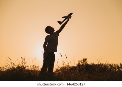 Black Silhouette Of Young Caucasian Kid Isolated On Sunny Golden Sunset Sky Background. Boy Playing Toy Plane Outside On Grassy Summer Hill. Setting Big Goals And Dreaming About Happy Future Concept.