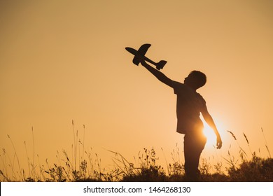 Black Silhouette Of Young Caucasian Kid Isolated On Sunny Golden Sunset Sky Background. Boy Playing Toy Plane Outside On Grassy Summer Hill. Setting Big Goals And Dreaming About Happy Future Concept.