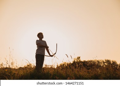 Black Silhouette Of Young Caucasian Kid Isolated On Sunny Golden Sunset Sky Background. Boy Playing Toy Bow And Arrow Outside On Summer Hill. Setting Big Goals Concept. Horizontal Color Photography.