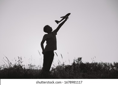 Black Silhouette Of Young Caucasian Kid Isolated On Sunny Sunset Sky Background. Boy Playing Toy Plane Outside On Grassy Summer Hill. Setting Big Goals And Dreaming About Happy Future Concept.