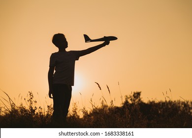 Black Silhouette Of Young Caucasian Kid Isolated On Sunny Golden Sunset Sky Background. Boy Playing Toy Plane Outside On Grassy Summer Hill. Setting Big Goals And Dreaming About Happy Future Concept.