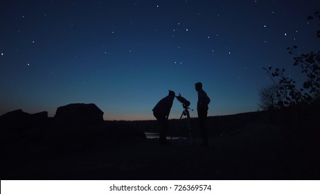 Black Silhouettes Of People Looking Through Telescope On Stars In Dark Twilight On Shore.