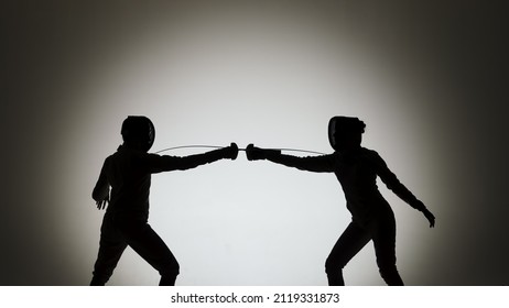 Black silhouettes of two female swordsmen fighting a duel. Athletes cross rapiers and deliver attacking blows. The girls pose in a dark studio on a white background in a spotlight spot. Slow motion. - Powered by Shutterstock