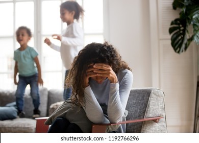 Black single frustrated woman hold her head with hands sitting on chair in living room, playful kids jumping on couch on a background. Tiredness, depression difficult to educate children alone concept