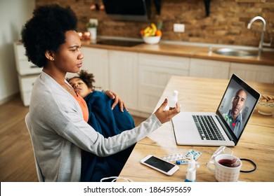 Black Single Mother Using Laptop And Having Online Appointment With A Doctor While Holding Her Sick Child In Her Lap At Home.