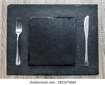 schwarze Schieferplatte und Schieferplatte mit Gabel und Messer auf einem Holztisch. Draufsicht einer Tischeinstellung