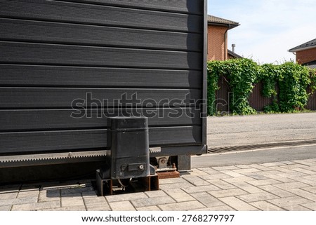 black sliding gate with an automatic drive mechanism, mounted on a paved area. Behind it a wooden fence covered with green ivy, adjacent to brick houses under a clear sky