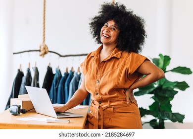 Black Small Business Owner Smiling At The Camera While Standing In Her Shop. Happy Businesswoman Managing Her Clothing Orders On A Laptop. Young Female Entrepreneur Running An Online Clothing Store.