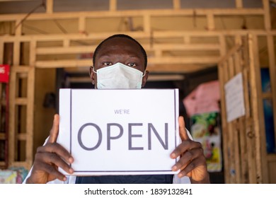 Black Small Business Owner Wearing A Face Mask And Holding An Open Sign In Front Of His Shop