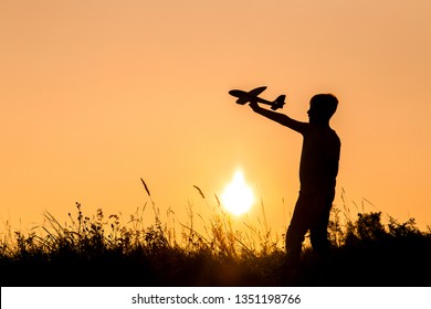 Black Small Silhouette Of Young Happy Kid Playing Toy Plane Outdoors At Sunset Time. Horizontal Color Photography.