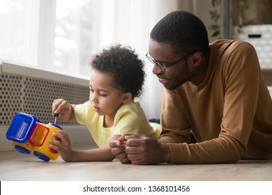 Black small son and father lying on warm floor play together loving dad looking at kid holds screwdriver repair fix toy truck car. Concept of teaching share experience playfully, developing creativity