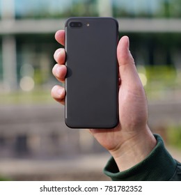 Black Smart Phone In A Man's Hand Back View On A Blurry City Background. Template Of Phone Case