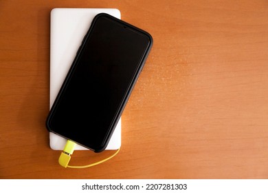 Black Smartphone Being Charged By A White Power Bank On Top Of A Wooden Table. Power Bank Concept. Technology Concept.