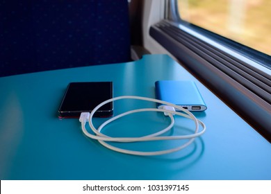 Black Smartphone Charging With Blue Power Bank On Blue Table Background Near The Seat Inside The Train Against Sunshine Through The Window.