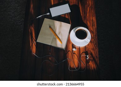 Black smartphone with light screen, paper notebook, pen, headphone and cup of coffee lies on a brown wooden table mahogany with spots of sunlight. Music and writter concept. Quarantine, self-isolation