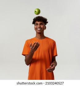 Black smiling man tossing up green organic apple. Concept of dieting and healthy eating. Young curly sporty guy wearing orange t-shirt. Isolated on white background. Studio shoot. Copy space - Shutterstock ID 2103240527
