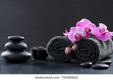 Black spa setting with grey towels, hot stones and beautiful orchids. Spa and wellness background with stack of hot stones with pink flowers on blackboard. Luxury spa composition and relax concept.