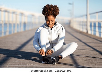 Black sportswoman tying shoelaces before running outdoors on wooden pier, sporty african american lady getting ready for jogging, enjoying healthy lifestyle and outside workouts, copy space - Powered by Shutterstock