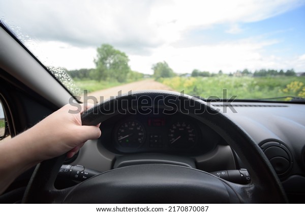 black steering wheel in a car from the first\
person on a road in a country with a cloudy sky, windshield wipers\
working, hand lying on a\
wheel