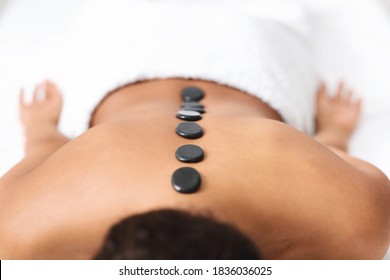 Black stones on naked unrecognizable black lady back, spa interior, closeup. Cropped of african american good looking woman attending spa salon, getting healing hot stone massage
