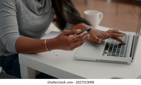 Black student holding cred card in hands doing electronic transcation searching online store, easy payment using digital gadget. Teenager browsing internet banking service on laptop in living room
