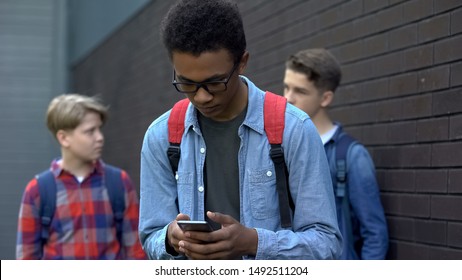 Black student reading offensive post in phone, boys mock behind, cyberbullying