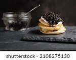 Black sturgeon caviar on small pancakes blinis with sour cream and a glass jar with caviar on a slate
