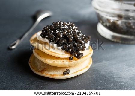 Black sturgeon caviar on small blini pancakes, a glass jar and a spoon on a dark gray background