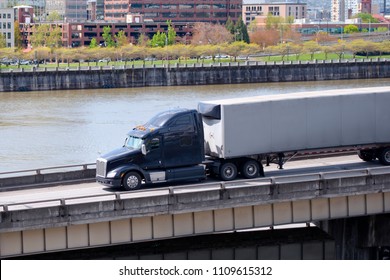 Black Stylish Big Rig American Semi Truck Tractor With Dry Van Semi Trailer Running On The Overpass Road Along The Willamette River In Portland City Limit For Long Haul Delivery Of Commercial Cargo